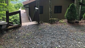 Eine Holzhütte mit einem Kiesweg, der zu einer Holzterrasse führt, umgeben von Bäumen und einem bewaldeten Hang.