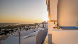 Un balcón con vistas a un pueblo costero al atardecer, una pequeña mesa con una botella y un vaso, y una alfombra azul.