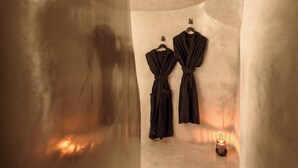Un pasillo estrecho y tenuemente iluminado con dos batas colgadas en ganchos en la pared, y una vela en el suelo.