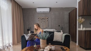Ein modernes Wohnzimmer mit einer Person, die eine Zeitschrift liest, einem runden Holztisch mit Dekorationsobjekten, einem grauen Sofa und einer Wand-Klimaanlage.