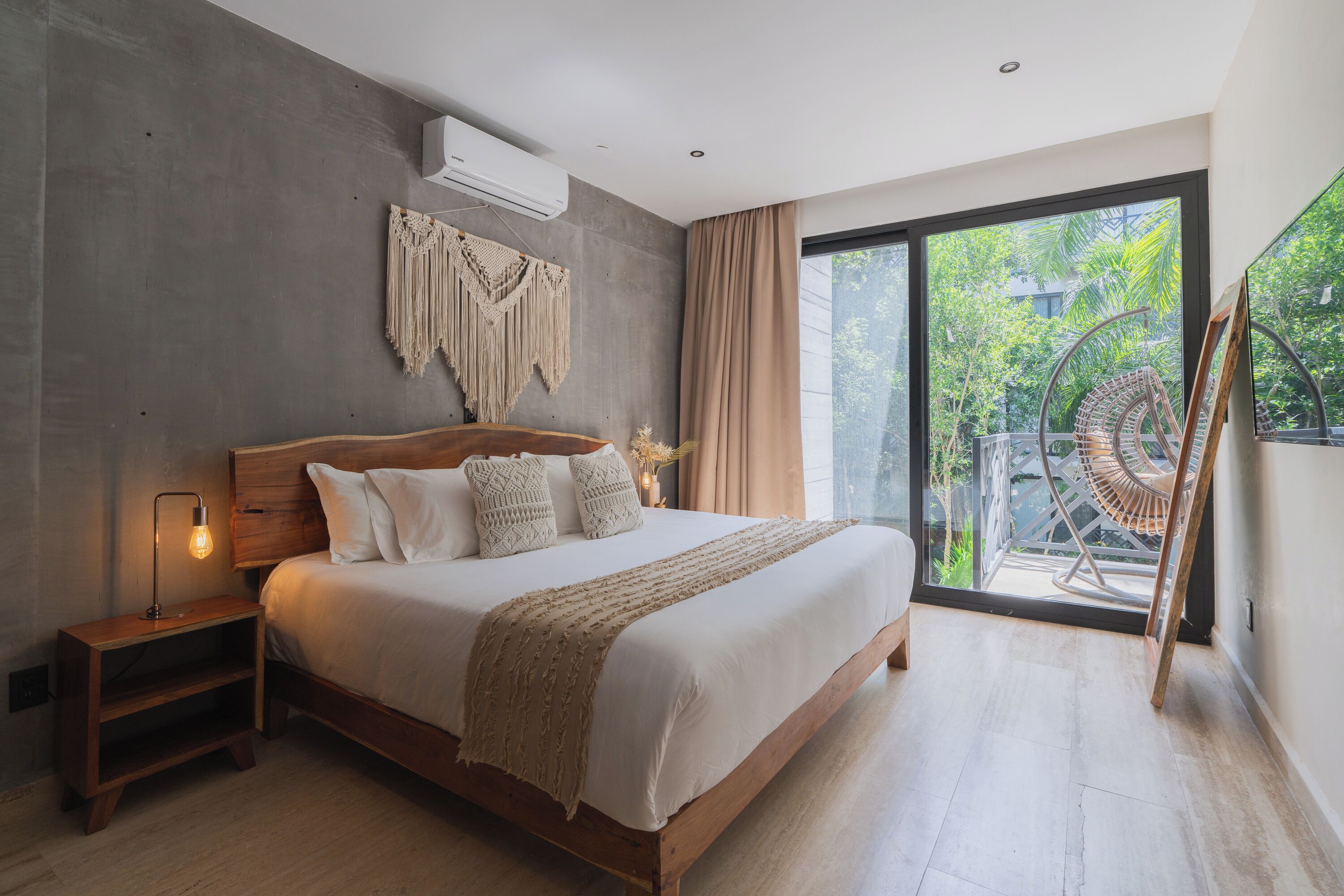 A modern bedroom with a large bed, a nightstand, a macrame wall hanging, and a view of greenery through a sliding glass door.