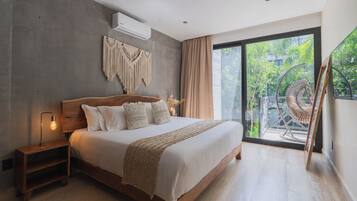 A modern bedroom with a large bed, a nightstand, a macrame wall hanging, and a view of greenery through a sliding glass door.