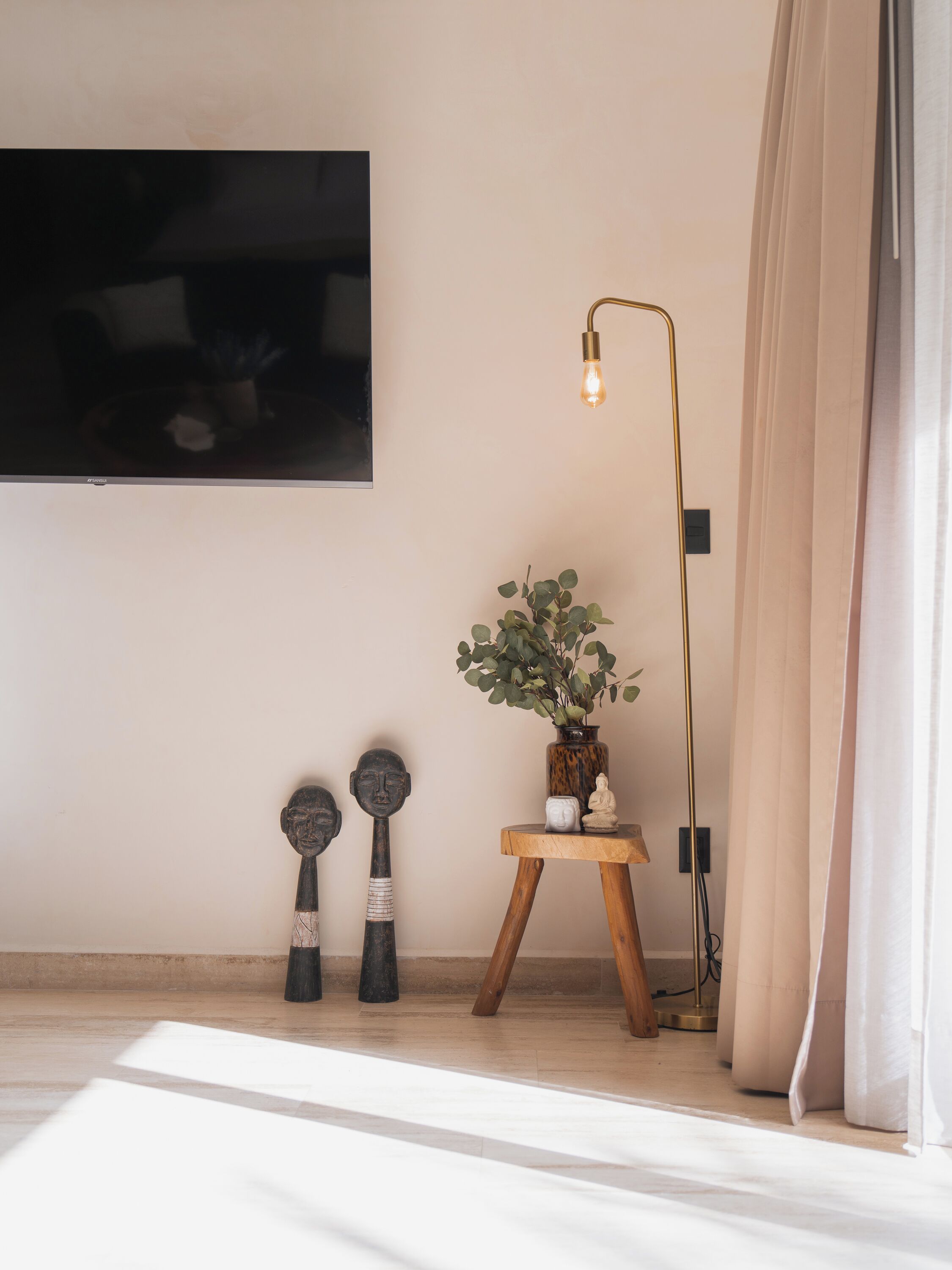 A room with a large flat-screen TV mounted on the wall, a floor lamp with a light bulb, a small wooden table with a vase of green plants, and two decorative wooden sculptures.