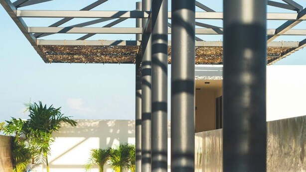 Ein moderner Poolbereich im Freien mit einer geometrischen Metallkonstruktion darüber, umgeben von Blumentöpfen und blauem Himmel.