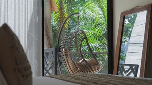Ein Korbstuhl mit Kissen, ein Bett mit strukturierter Decke und Blick auf Palmen durch eine Glastür.
