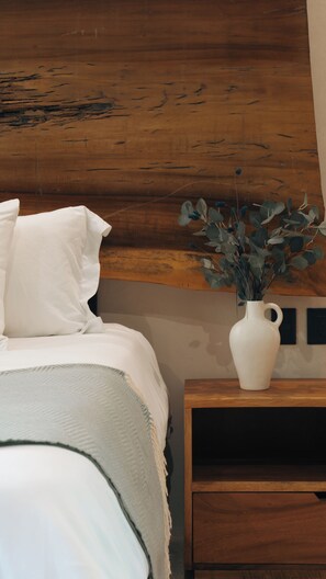A bedroom with a wooden headboard, a white bed, and a nightstand with a white vase containing green foliage.