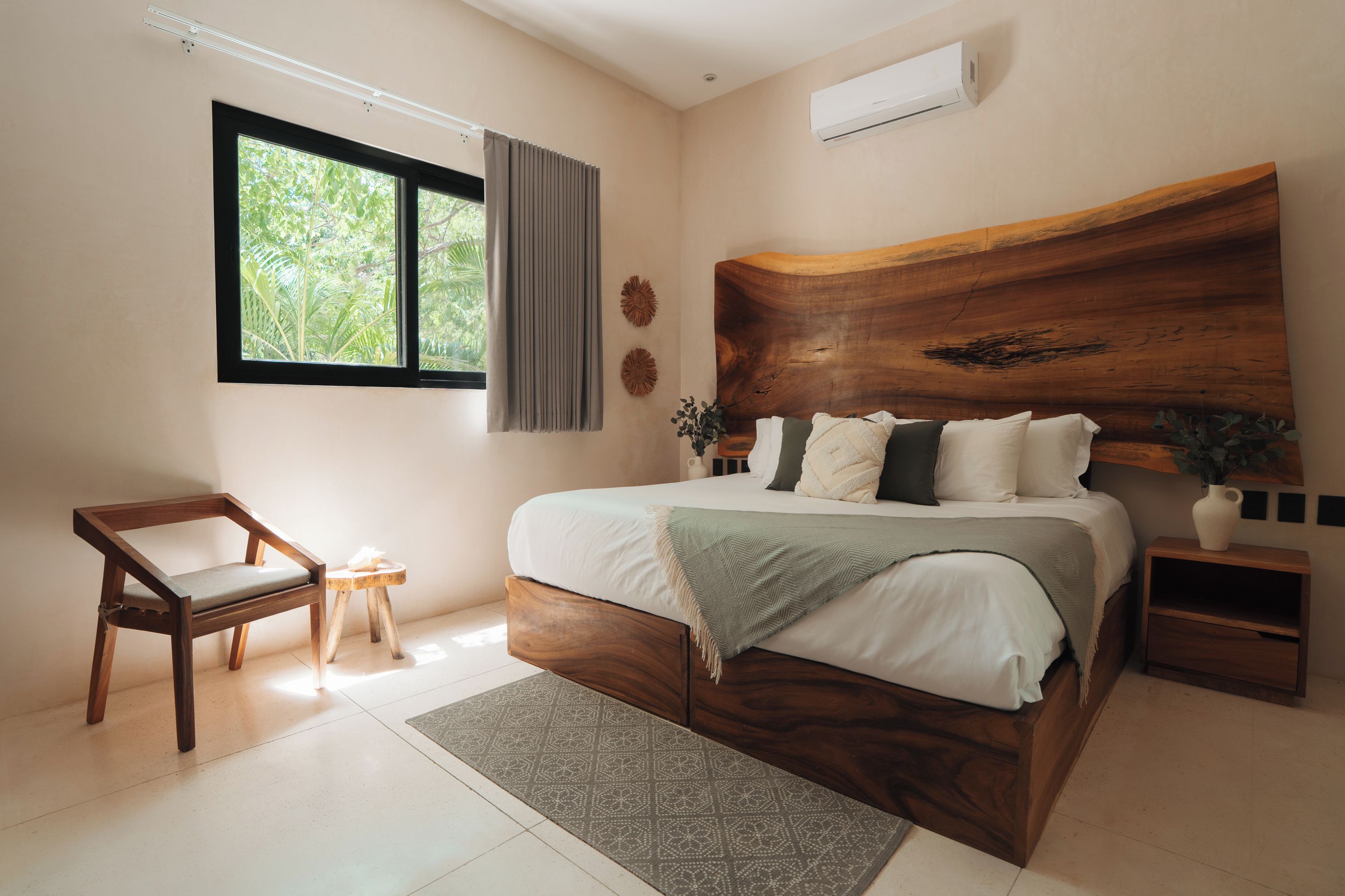 Ein Schlafzimmer mit hölzernem Kopfteil, einem Bett mit weißer Bettwäsche, einem Holzsessel, einem kleinen Nachttisch und einem Fenster mit Vorhängen.