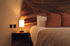 Cama bem arrumada com travesseiro branco e dois travesseiros estampados, luminária de cabeceira em uma mesinha de madeira, e parede com design em painel de madeira.
