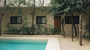 A pool area with a clear blue pool, surrounded by trees and plants, and a building with windows and a wooden door.