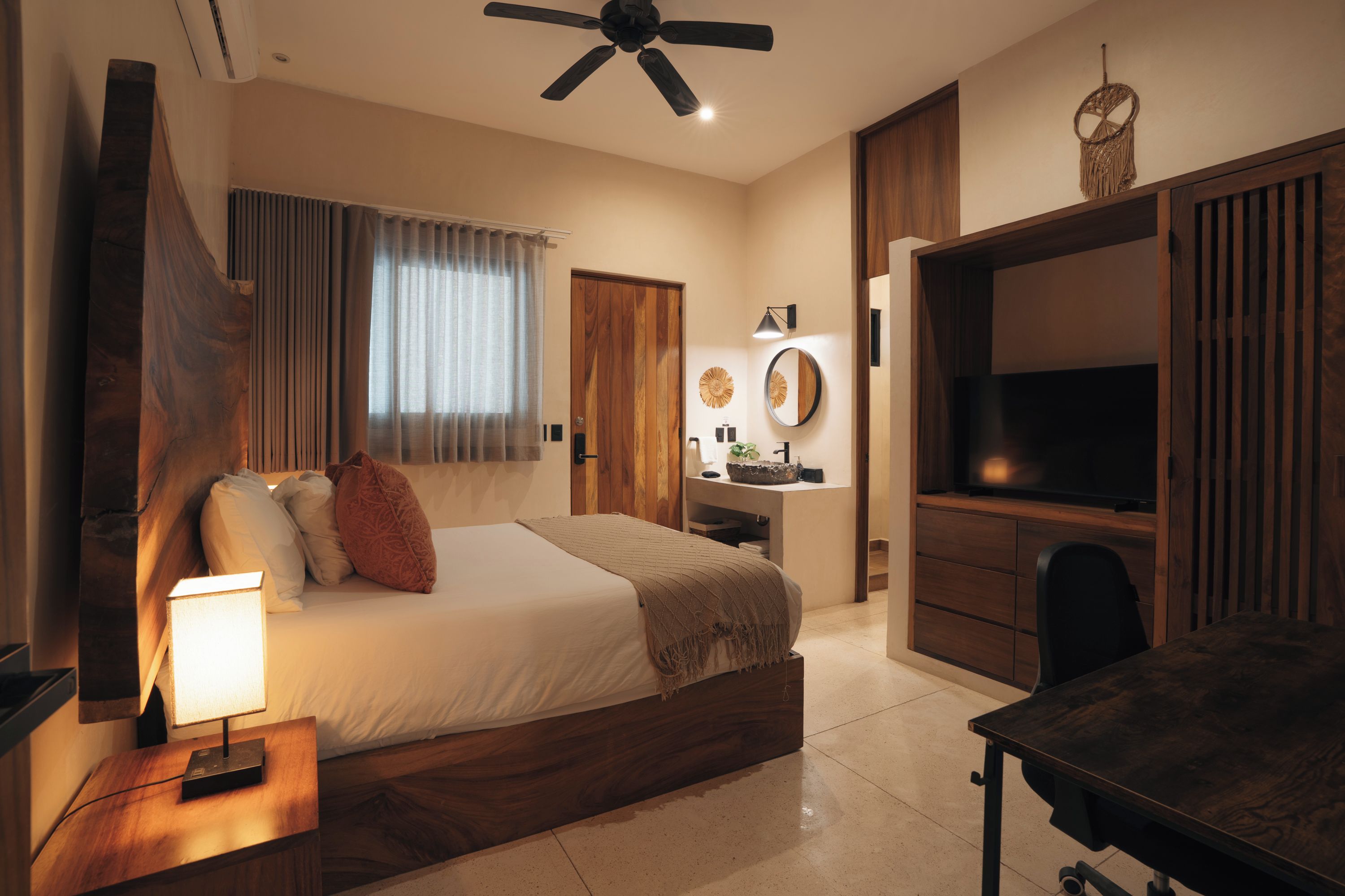 Habitación de hotel con cama, mesita de noche, lámpara, ventilador de techo, televisor y escritorio con espejo.