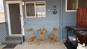 Un patio couvert avec deux chaises en bois, une petite table, un barbecue et une bouteille de gaz.