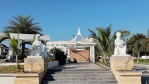 A pathway leading to a building with a distinctive roof, flanked by statues and palm trees. - The Grand Rudraksh (Amravati)