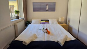 A hotel room with a bed, a nightstand, a lamp, a window with curtains, a potted plant, and a painting of a lighthouse.