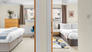 A hotel room with two beds, a desk, a chair, a radiator, and a view of the street.