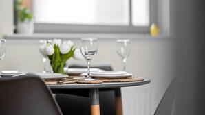 A dining table with a vase of white flowers, clear glasses, and plates, set in a modern room with a window and a plant in the background.