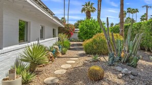 A house with a garden pathway, cacti, palm trees, and a clear sky.
