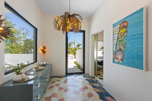 Un pasillo moderno con un suelo de baldosas hexagonales de colores, un gabinete de vidrio con objetos decorativos, una lámpara colgante de forma de hoja original y un cuadro abstracto grande en la pared.