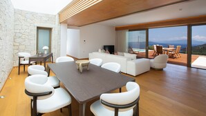 A modern living room with a dining area, stone walls, wooden flooring, and large windows offering a view of the outdoors.