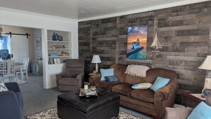 Un salón con un sofá marrón, un otoman negro y un sofá color beige. Se aprecia una pared de madera con una pintura de atardecer y una decoración náutica.
