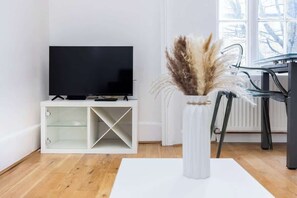 A modern living room with a flat-screen TV on a white console, a glass vase with dried plants, and a dining area with a black chair.