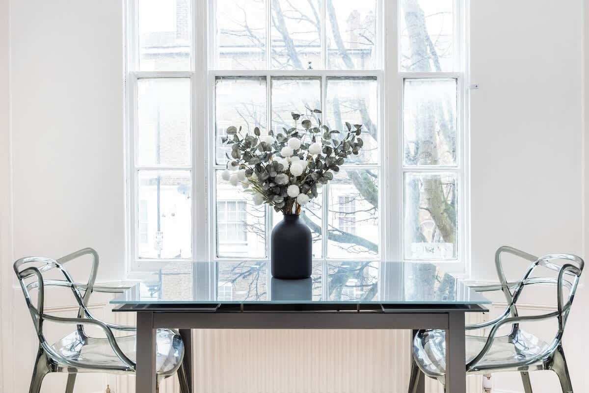 Une pièce avec une table et des chaises en verre, un vase rempli de fleurs et une grande fenêtre donnant sur des immeubles.