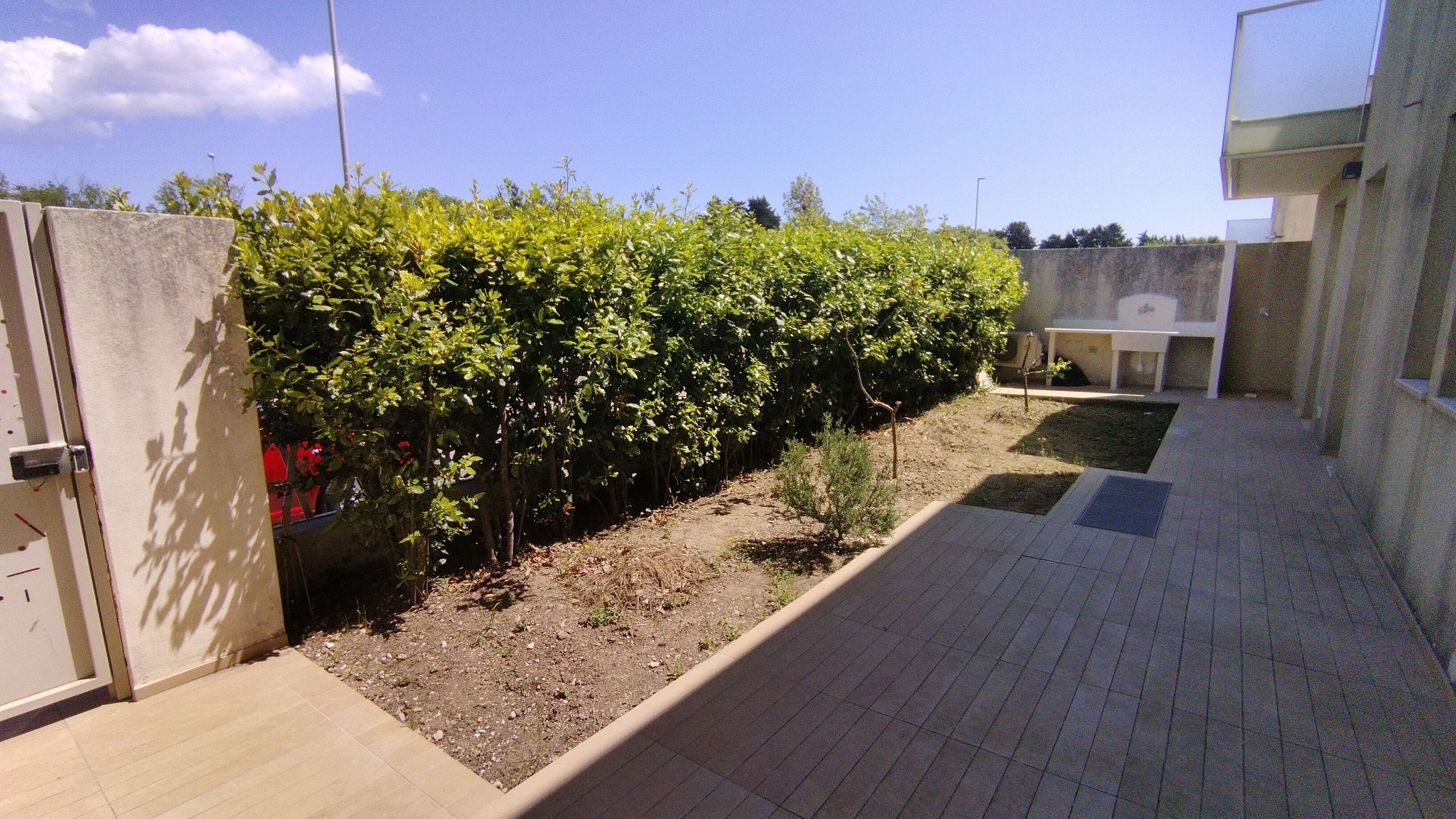 A paved outdoor area with a hedge, a small garden bed, and a building with a flat roof.