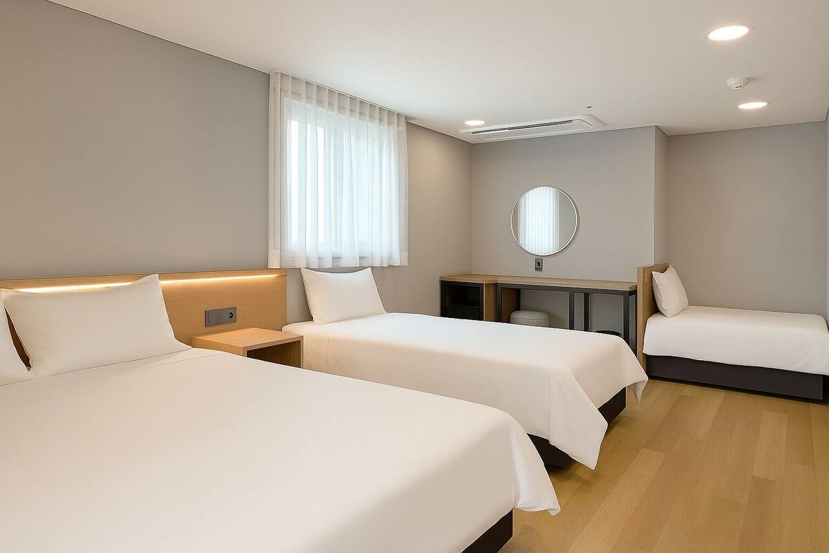 A hotel room with three beds, a desk, a mirror, and a window with sheer curtains.