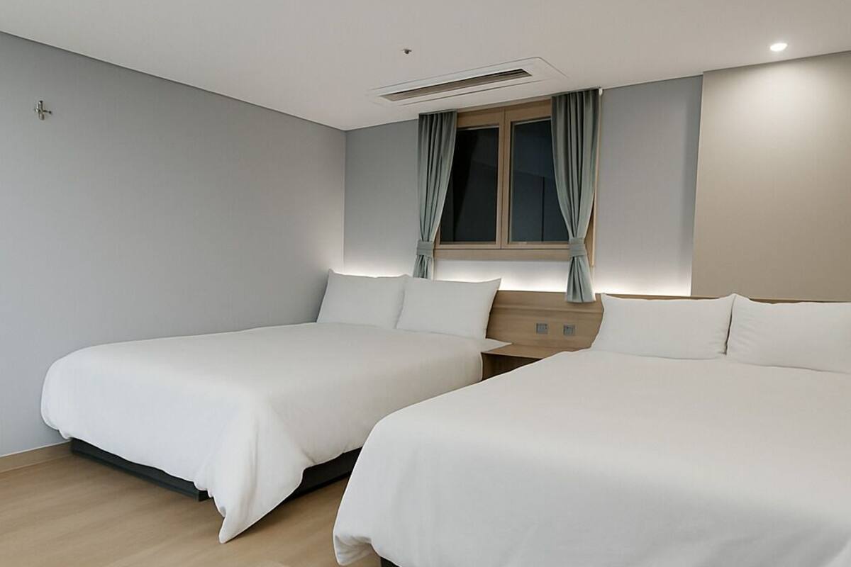 A modern hotel room with two single beds, a window with curtains, a wooden floor, and a ceiling light.