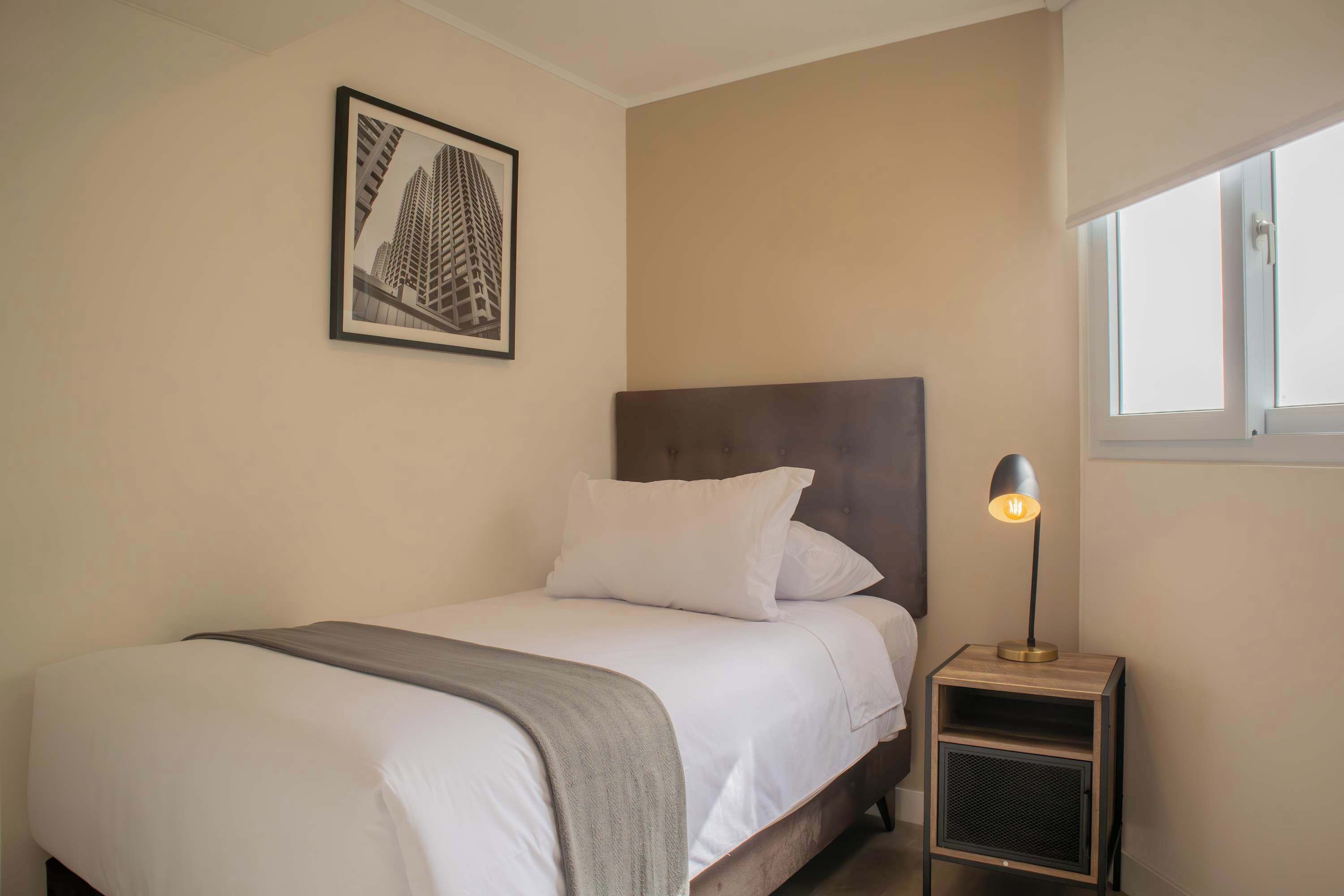 A neatly made bed with white linens and a grey throw blanket, a framed picture on the wall, a bedside lamp, and a window with a view of the outside.