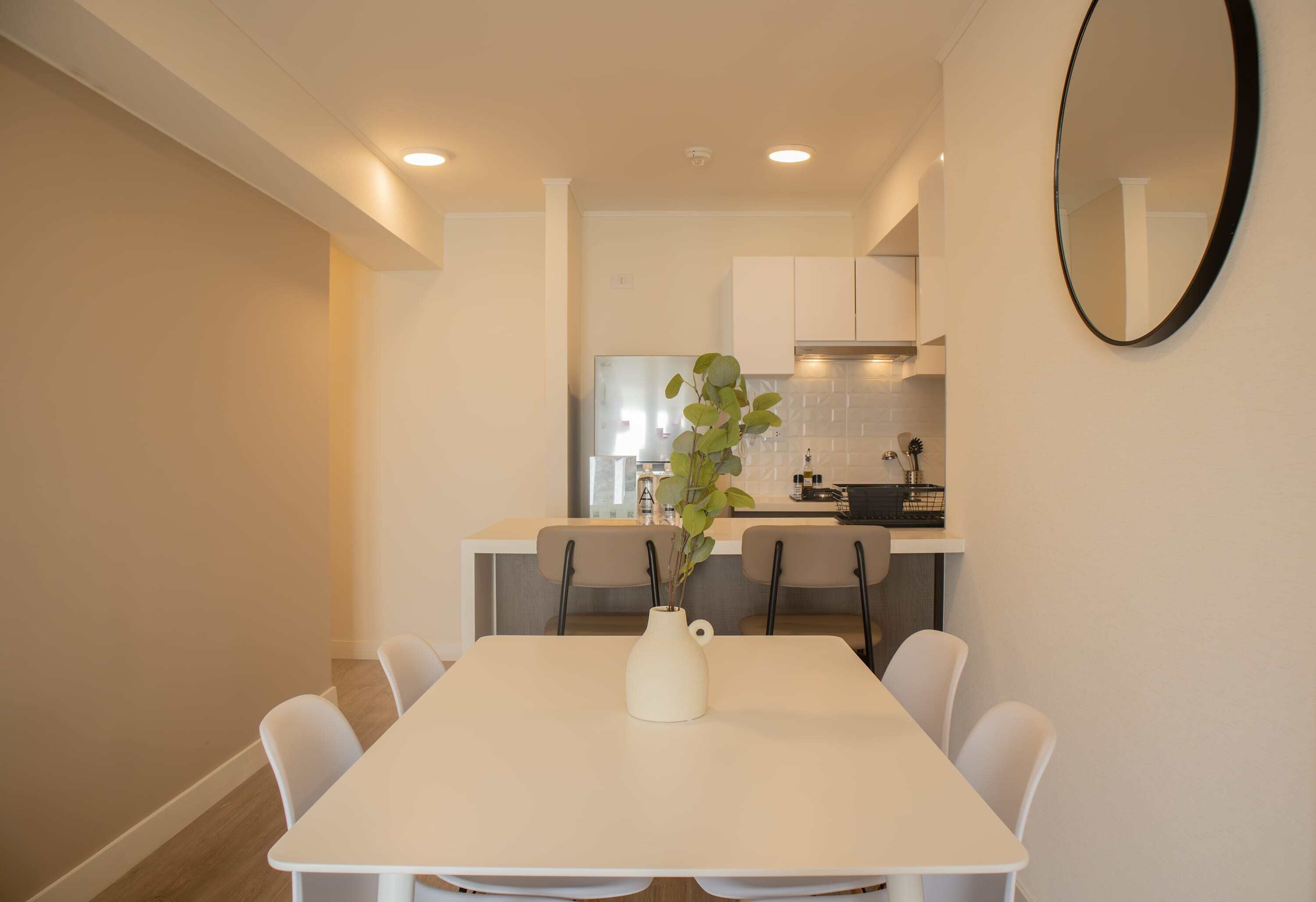 Un comedor con una mesa y sillas blancas, un jarrón con planta y una cocina al fondo.