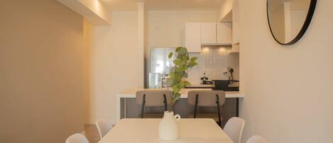 Un comedor con una mesa y sillas blancas, un jarrón con planta y una cocina al fondo.