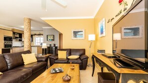 Ein Wohnzimmer mit braunem Ledersofa, einem Holztisch und einer TV-Konsole. Im Hintergrund befindet sich eine Küche mit einer gefliesten Spritzschutzwand und ein Essbereich mit Tisch und Stühlen.