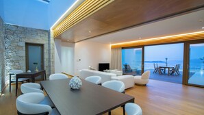 A modern living room with a stone accent wall, a dining area with a wooden table and white chairs, and a large window offering a view of the sea. - 2S Villas Resort - cave villas - Villa Epsilon E - Luxury Comfort Stay (Sivota)