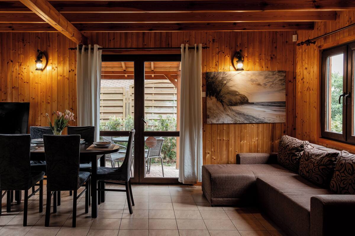 A wooden interior with a dining area, a sofa, and a large window with a beach view.