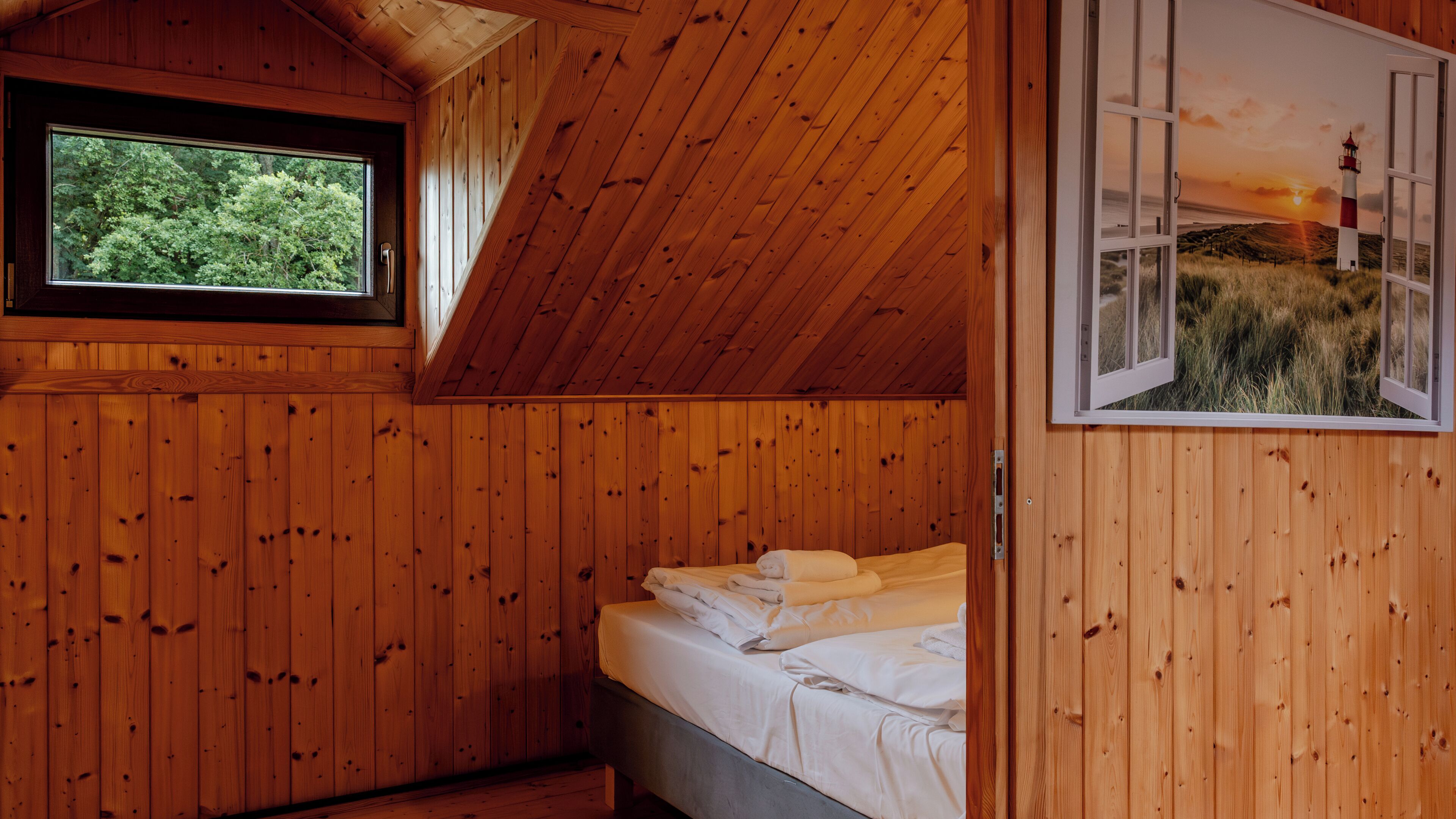 A wooden cabin room with a bed, a window, and a painting of a lighthouse.