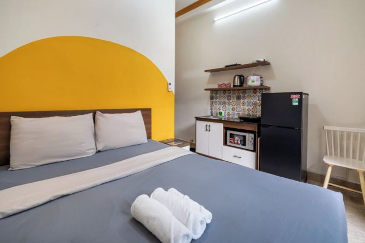 A hotel room with a bed, a small kitchenette, and a white chair.