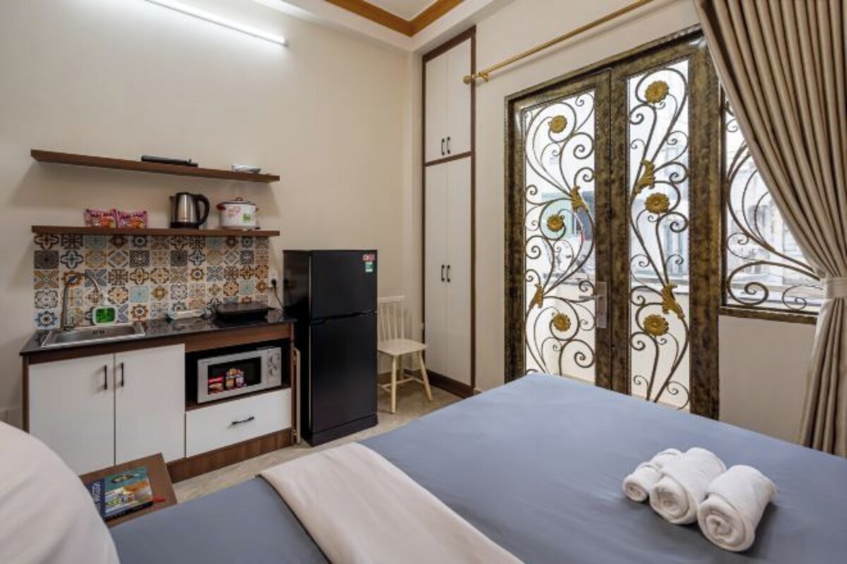 A hotel room with a bed, a small kitchenette, a decorative window, and a view of the city.