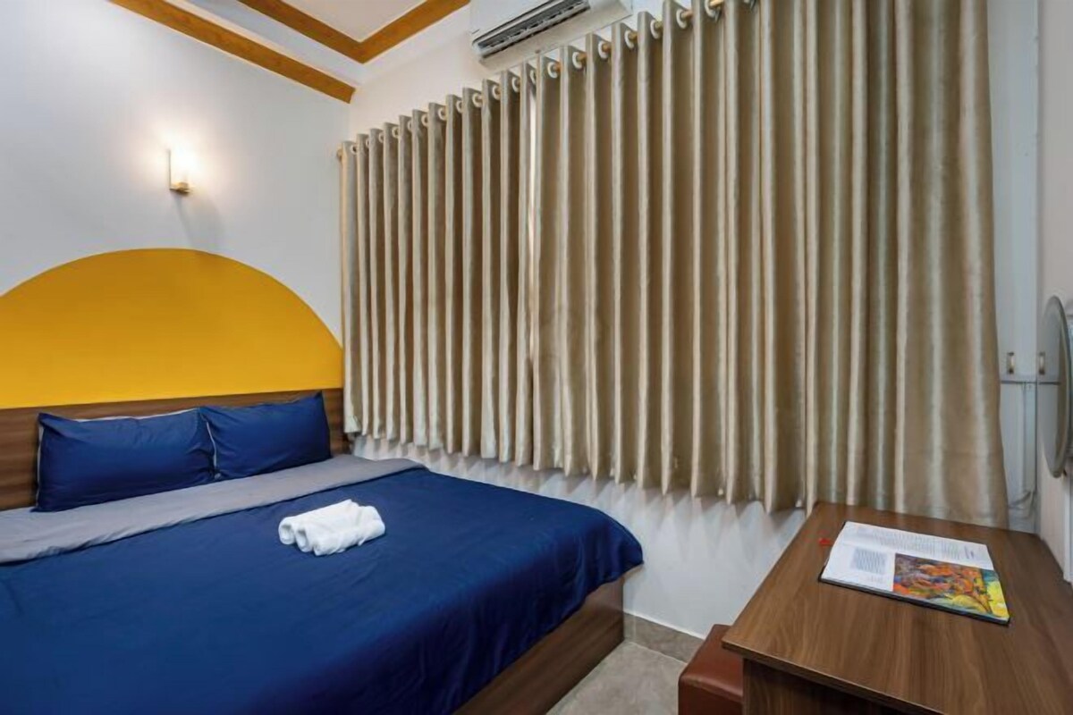 A hotel room with a bed, a wooden headboard, a window with beige curtains, a bedside table with a magazine, and a wall-mounted air conditioning unit.