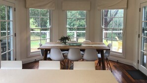 Un comedor con mesa de madera, sillas blancas y ventanas grandes con cortinas.