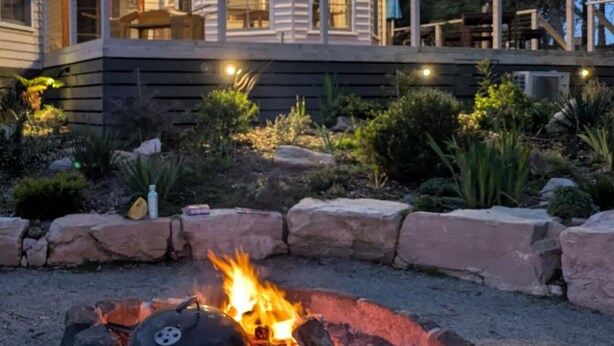 Una casa con un espacio al aire libre cubierto, una fogata y jardines a su alrededor.