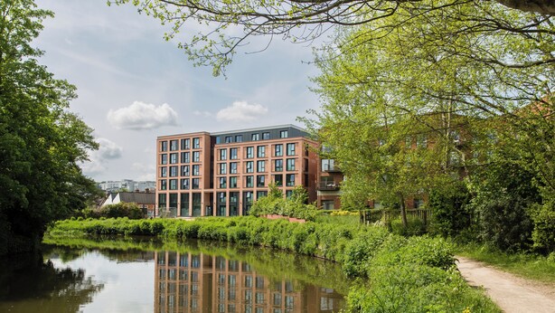 Un edificio moderno con facciata in mattoni e ampie finestre, adiacente a un fiume tranquillo e rigogliosa vegetazione.