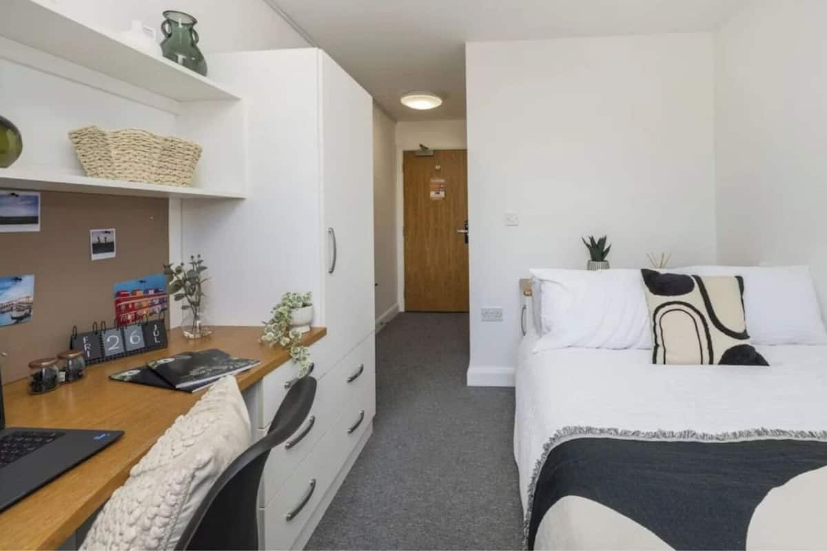 A small, clean room with a bed, a desk, and a chair. There are shelves with decorative items and a calendar on the wall.