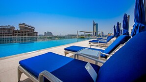 Un área de piscina en la azotea con sillas reclinables y sombrillas azules, con vistas a un paisaje urbano con edificios modernos.