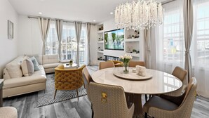 En moderne stue med sofa, spisebord, stole, en lysekrone og et fjernsyn på en reol.
