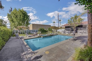 A swimming pool with lounge chairs and umbrellas, surrounded by greenery and a building.