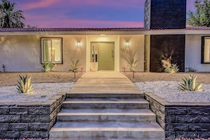 A modern house with a concrete staircase leading to a door, surrounded by landscaped areas with agave plants.