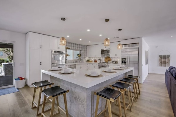 A modern kitchen with a central island, bar stools, stainless steel appliances, pendant lighting, and a view of the outdoors.