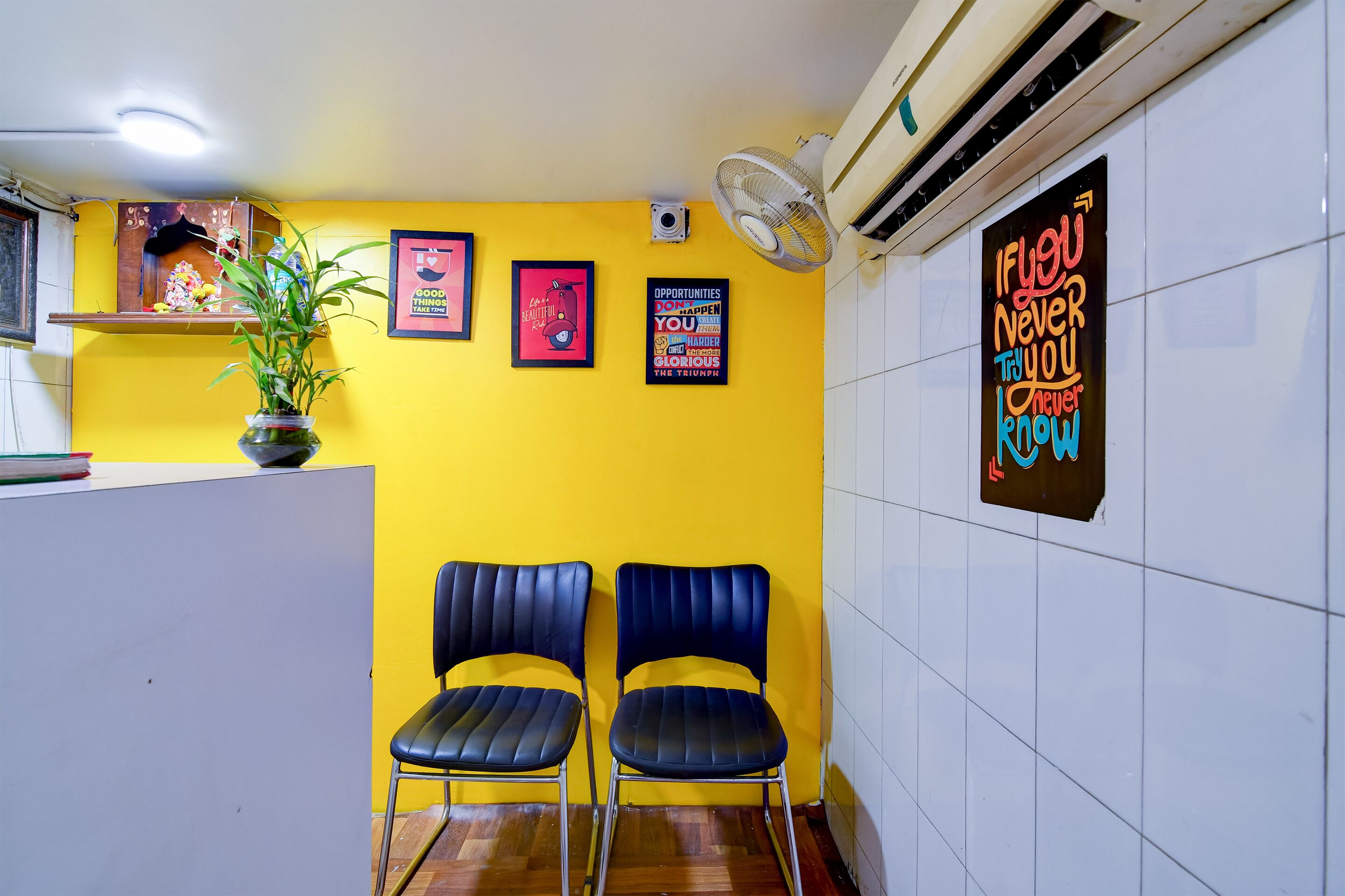 A reception area with a yellow wall, two blue chairs, a white reception desk, a potted plant, and motivational posters.