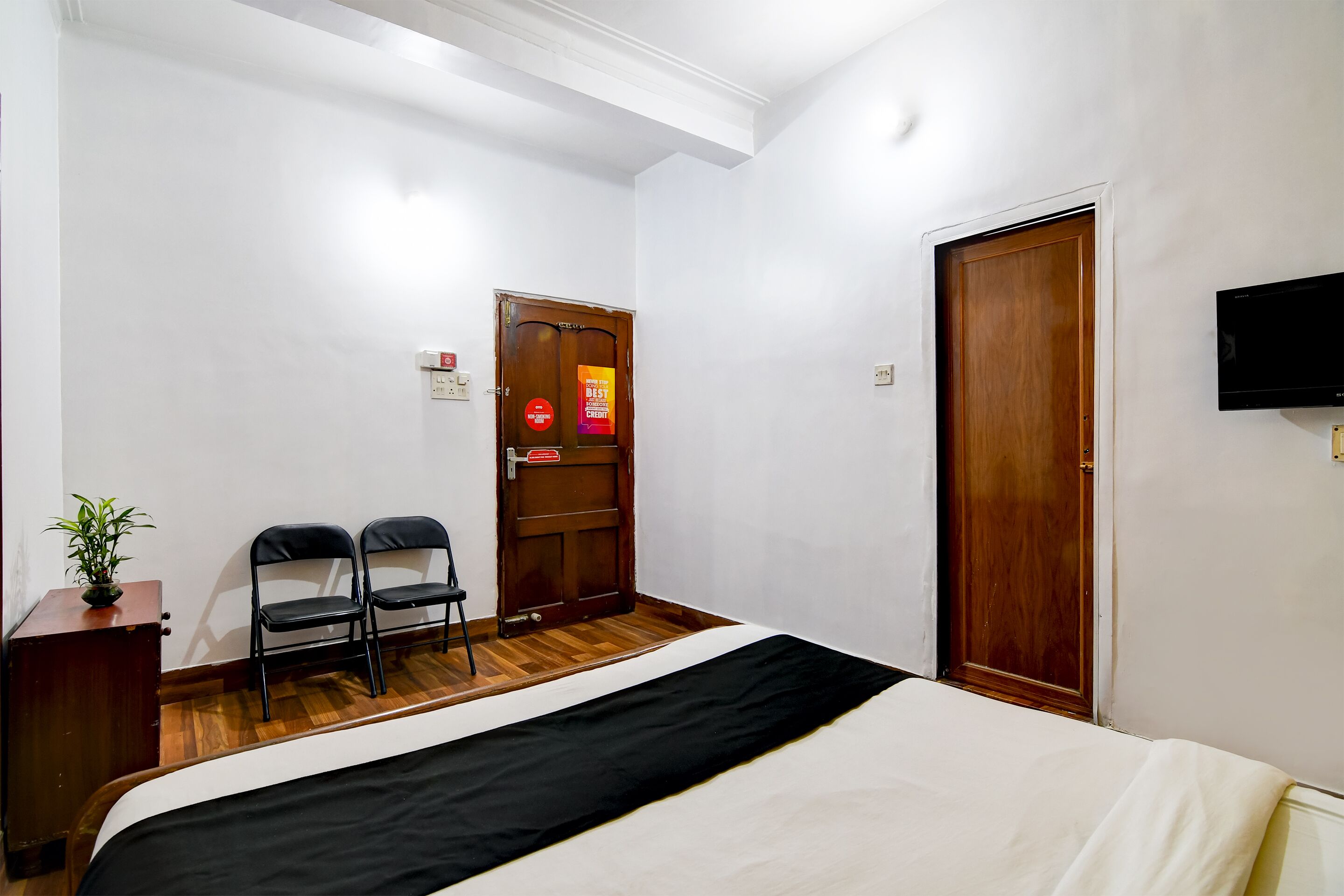A hotel room with a bed, two chairs, a wooden door, and a television on the wall.