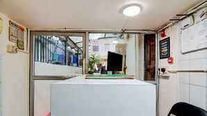 Een kamer met een bureau, een stoel, een raam met metalen tralies, een plant en diverse wanddecoraties, waaronder een poster en een ingelijst diploma.